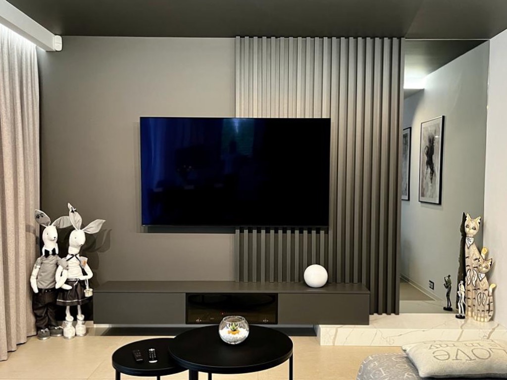 Nowoczesny salon w odcieniach szarości z telewizorem na ścianie, dekoracyjnymi lamelami, dwoma pluszowymi królikami i czarnymi stolikami kawowymi.