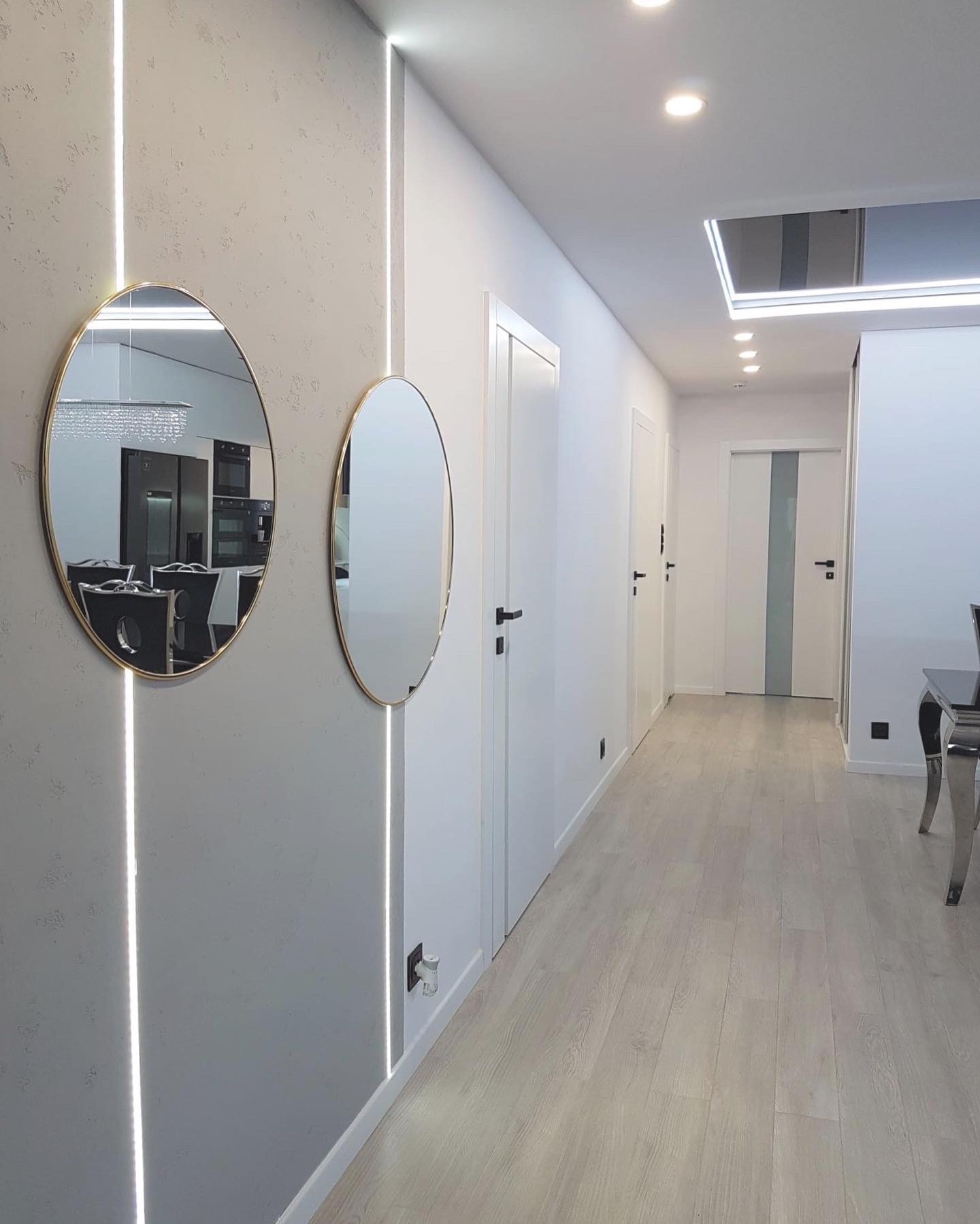 Nowoczesny korytarz z szarą teksturowaną ścianą, dwoma owalnymi lustrami w złotych ramach, pionowym oświetleniem LED, białymi drzwiami i jasną drewnianą podłogą.