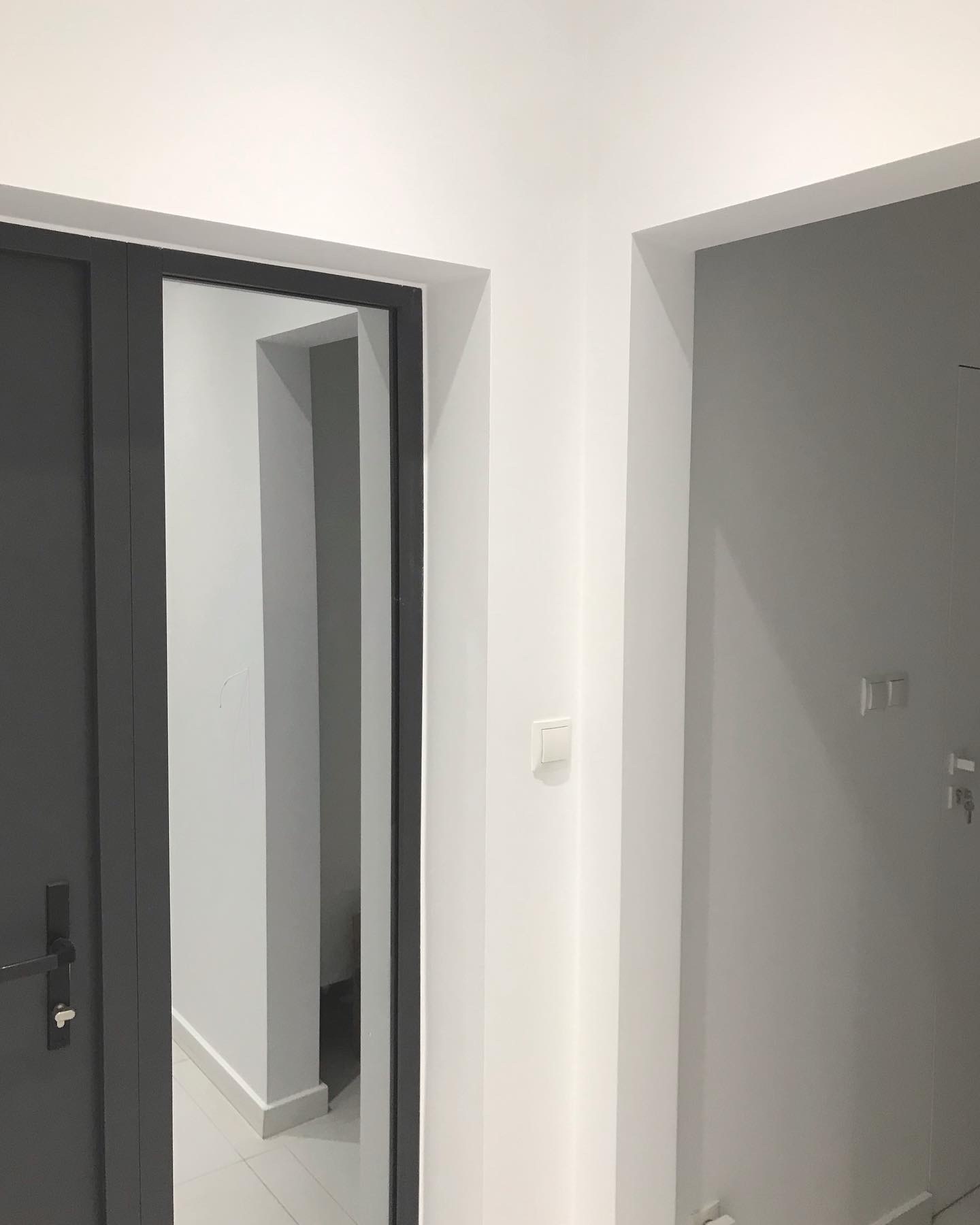 Wnętrze w odcieniach szarości i bieli: widok na korytarz przez otwarte, ciemnoszare drzwi, z widocznym włącznikiem światła na białej ścianie i kluczami w zamku szarych drzwi w tle.