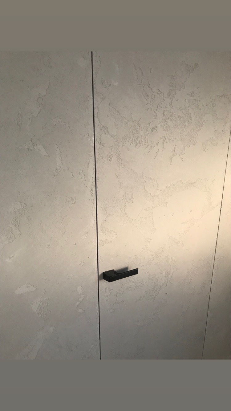 Ukryte drzwi w ścianie z efektem dekoracyjnym w odcieniach szarości, z czarną klamką, zrealizowane w Głogowie.