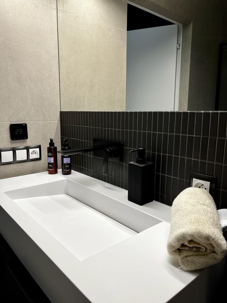 Nowoczesna łazienka z białą, prostokątną umywalką, czarną armaturą i pionową mozaiką na ścianie, uzupełniona o dozownik mydła w identycznym kolorze i zwinięty, kremowy ręcznik.