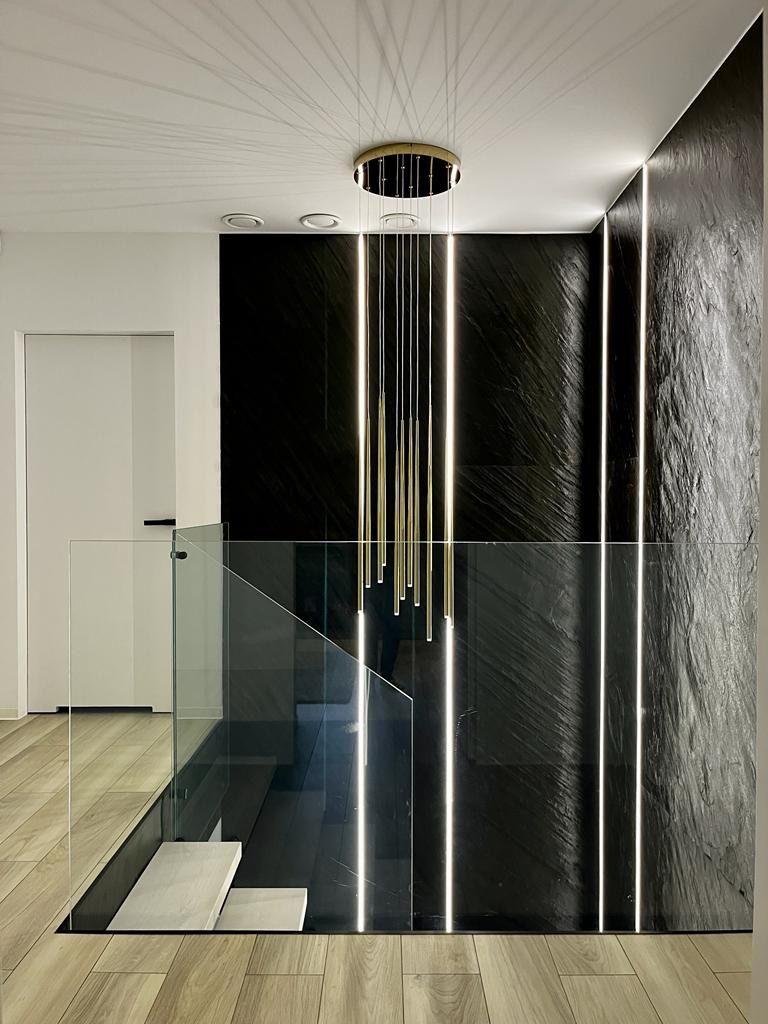 Nowoczesne wnętrze z podświetlaną ścianą z czarnego kamienia, szklaną balustradą schodów i designerską lampą wiszącą z cienkimi, złotymi prętami.