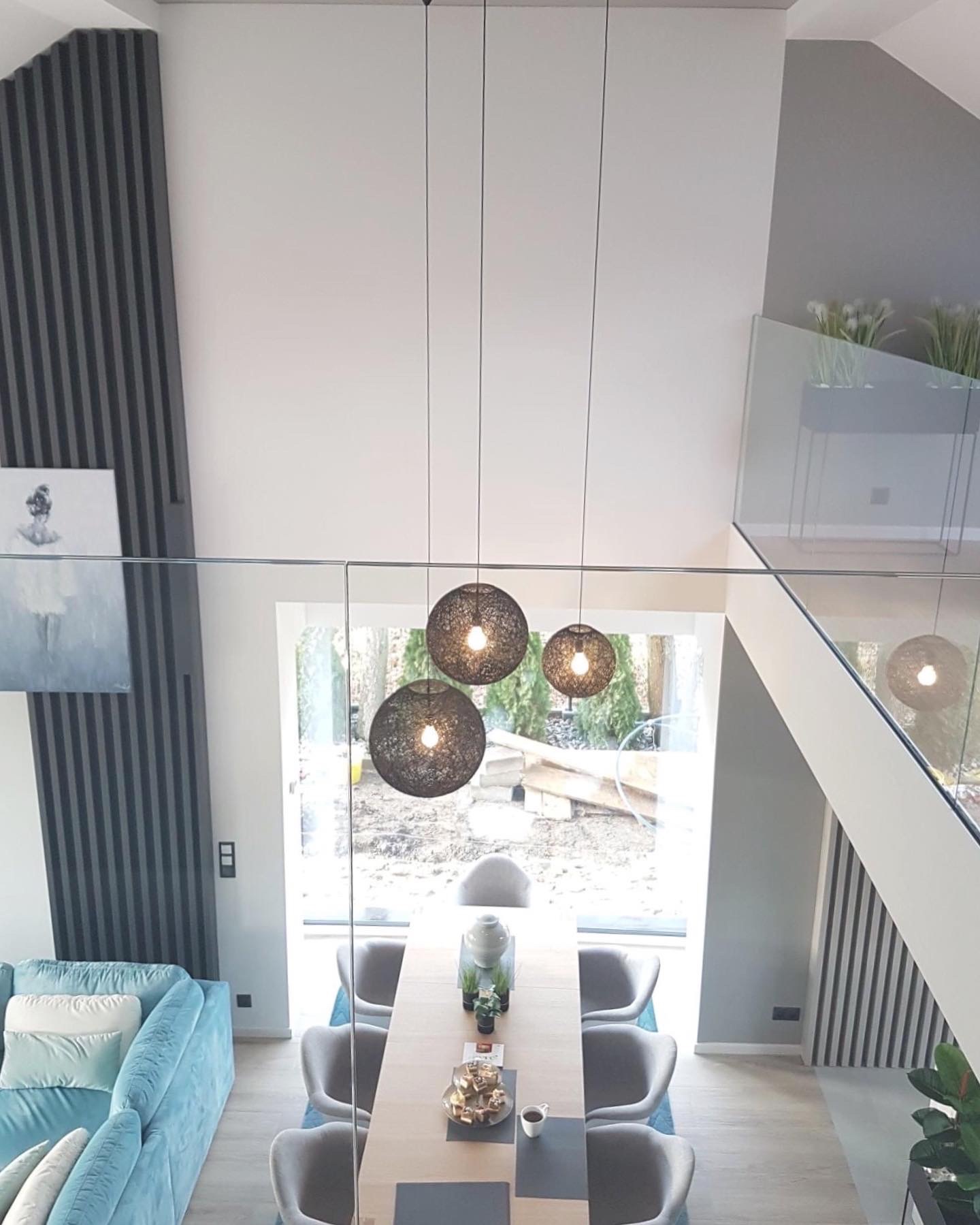 Dwupoziomowy salon z wysokim sufitem, wiszącymi lampami kulistymi, turkusową sofą i szklaną balustradą, widok z góry na stół jadalny i krzesła.