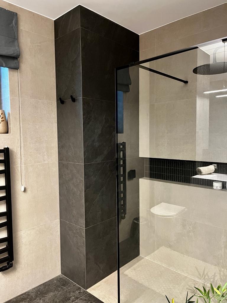 Nowoczesna łazienka z kabiną prysznicową typu walk-in, wykończona płytkami w odcieniach beżu i grafitu, z czarnymi akcentami w postaci ramy kabiny, grzejnika drabinkowego i haczyków na ścianie.