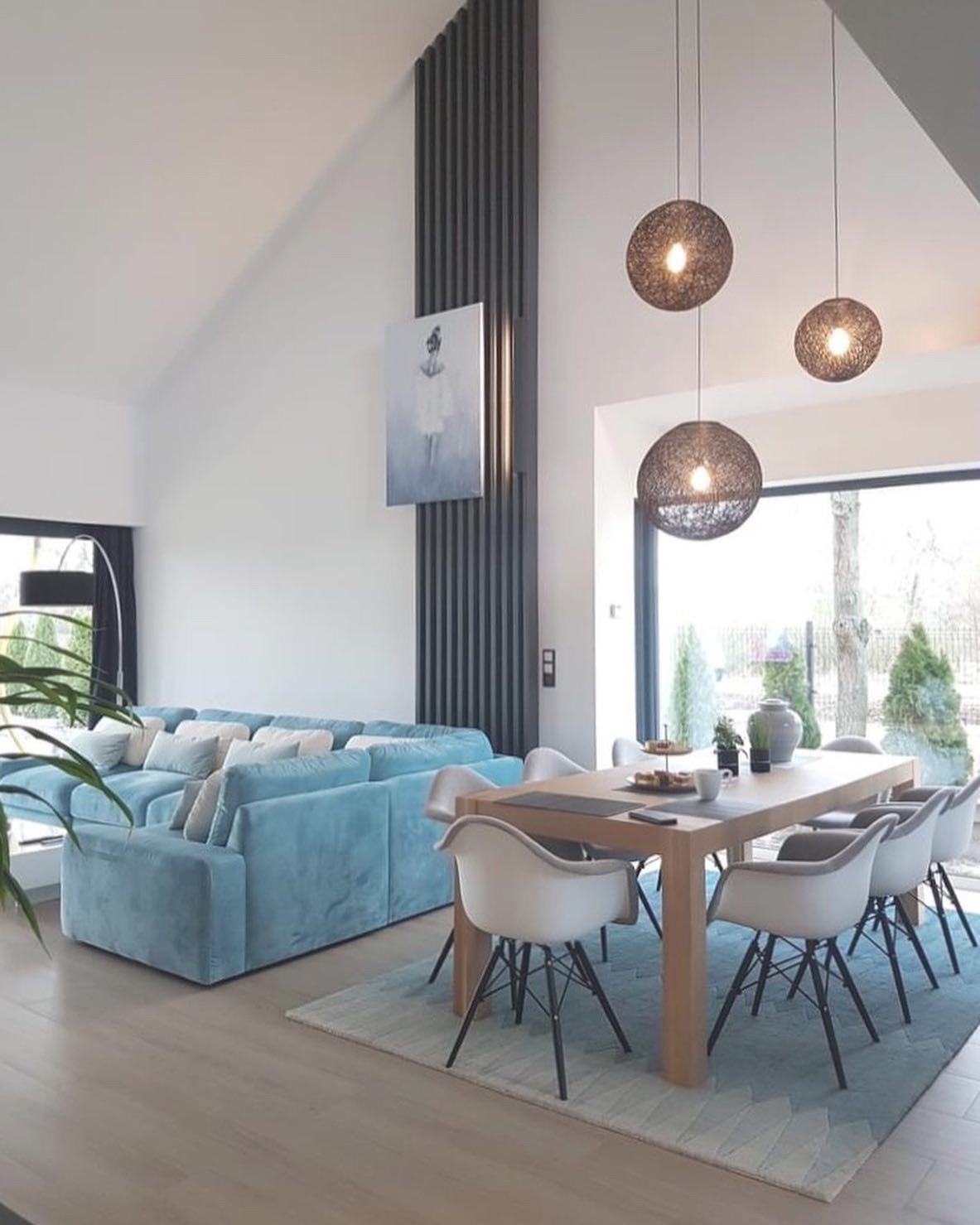 Jasny salon z wysokim sufitem, turkusową sofą, drewnianym stołem jadalnym z białymi krzesłami i trzema okrągłymi lampami wiszącymi, dekorowany ciemnoszarymi pionowymi listwami na ścianie i obrazem...