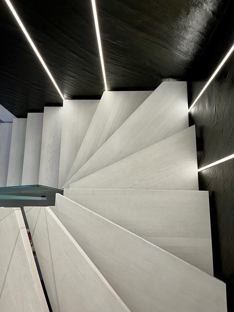 Nowoczesne, spiralne schody z jasnego drewna z podświetleniem LED na czarnej ścianie o fakturze kamienia.