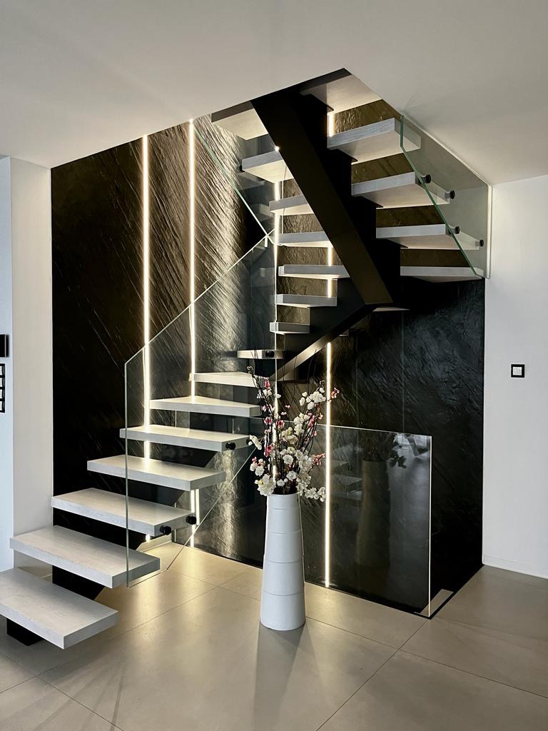 Nowoczesne schody wspornikowe z szarymi stopniami, czarną konstrukcją i szklanymi balustradami na tle ściany z czarnego kamienia z pionowym oświetleniem LED, wazon z kwiatami jako element dekoracyjny.
