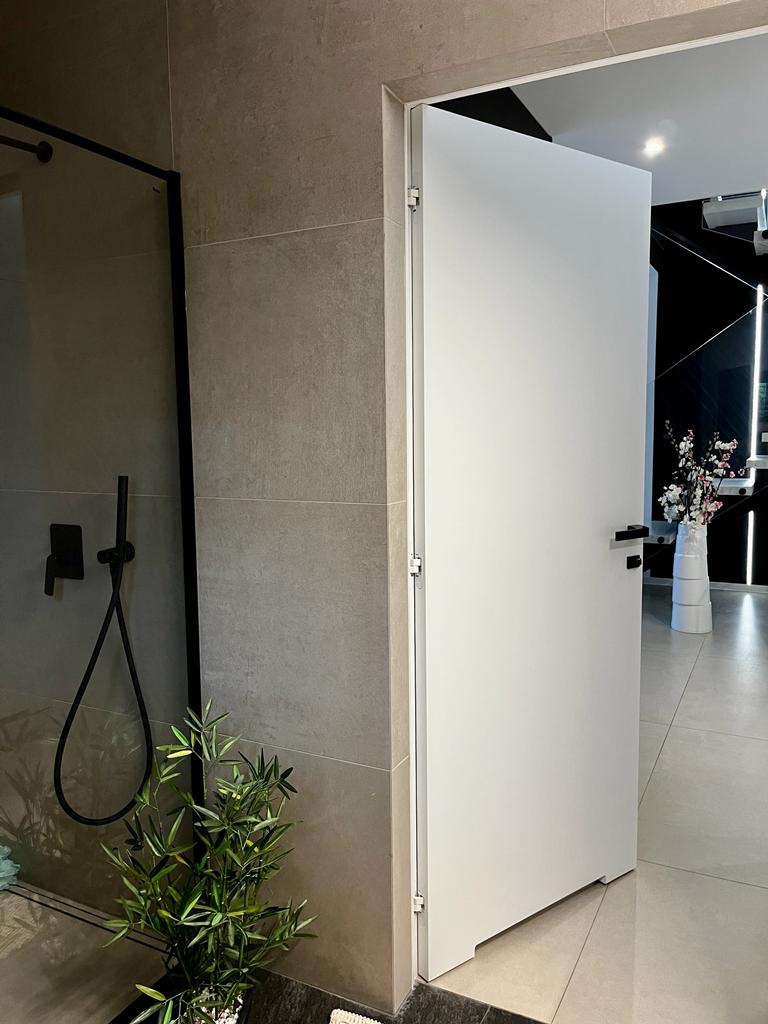 Nowoczesna łazienka z kabiną prysznicową typu walk-in, szarą glazurą i białą, minimalistyczną armaturą oraz drzwiami, z widoczną rośliną doniczkową w rogu.