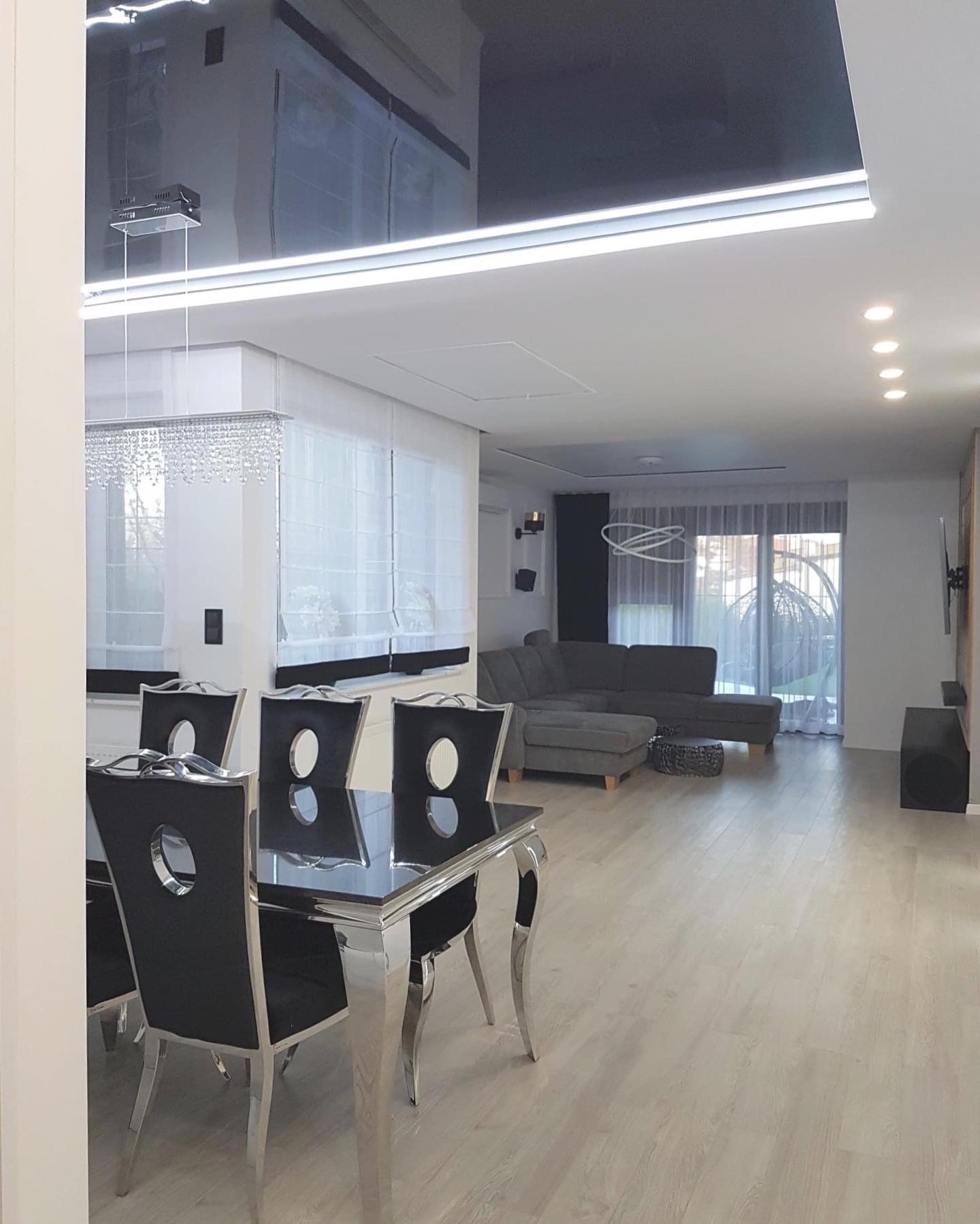 Nowoczesne wnętrze: czarny, lustrzany sufit napinany z oświetleniem LED, stół z czarnym blatem i chromowanymi nogami, krzesła z czarną tapicerką i chromowanymi detalami, szara sofa w tle.