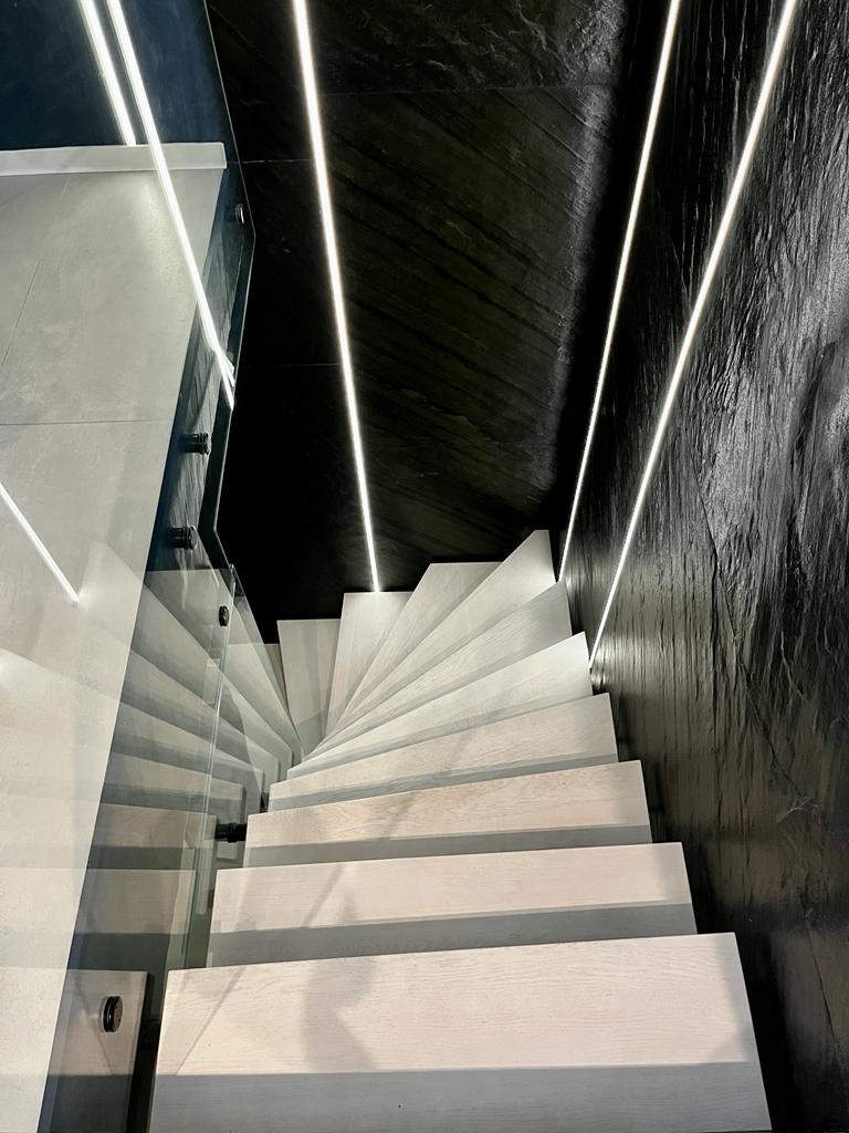 Spiralne schody z białego drewna z szklaną balustradą, oświetlone ledowymi paskami na czarnym, kamiennym tle ściany.
