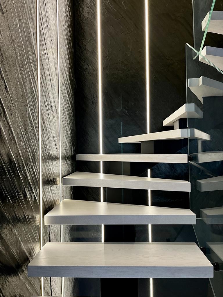 Nowoczesne schody z drewnianymi stopniami, szklaną balustradą i oświetleniem LED na ścianie o teksturze kamienia.