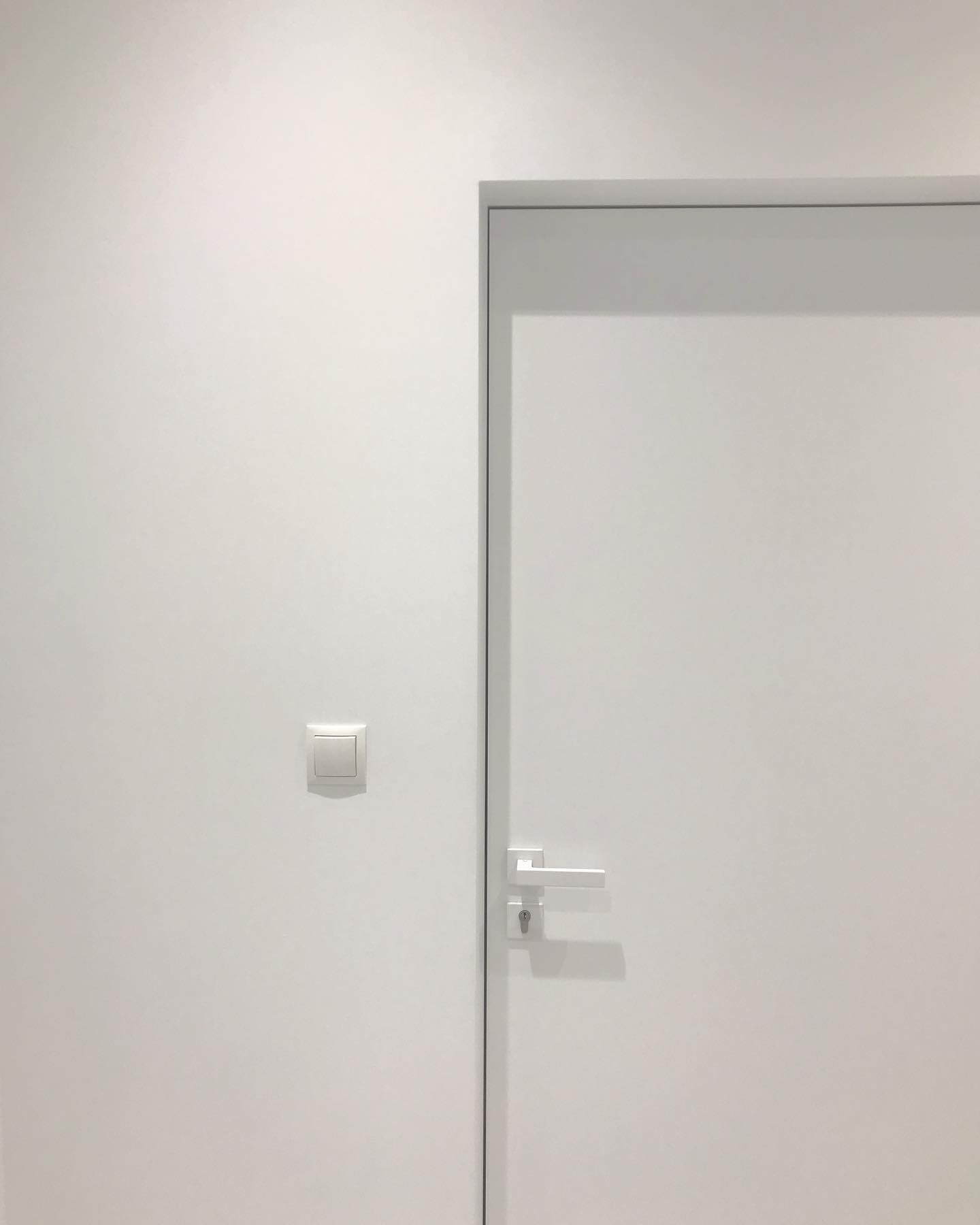 Minimalistyczne wnętrze: biała ściana z kwadratowym włącznikiem światła, białe drzwi z prostą, srebrną klamką i zamkiem.