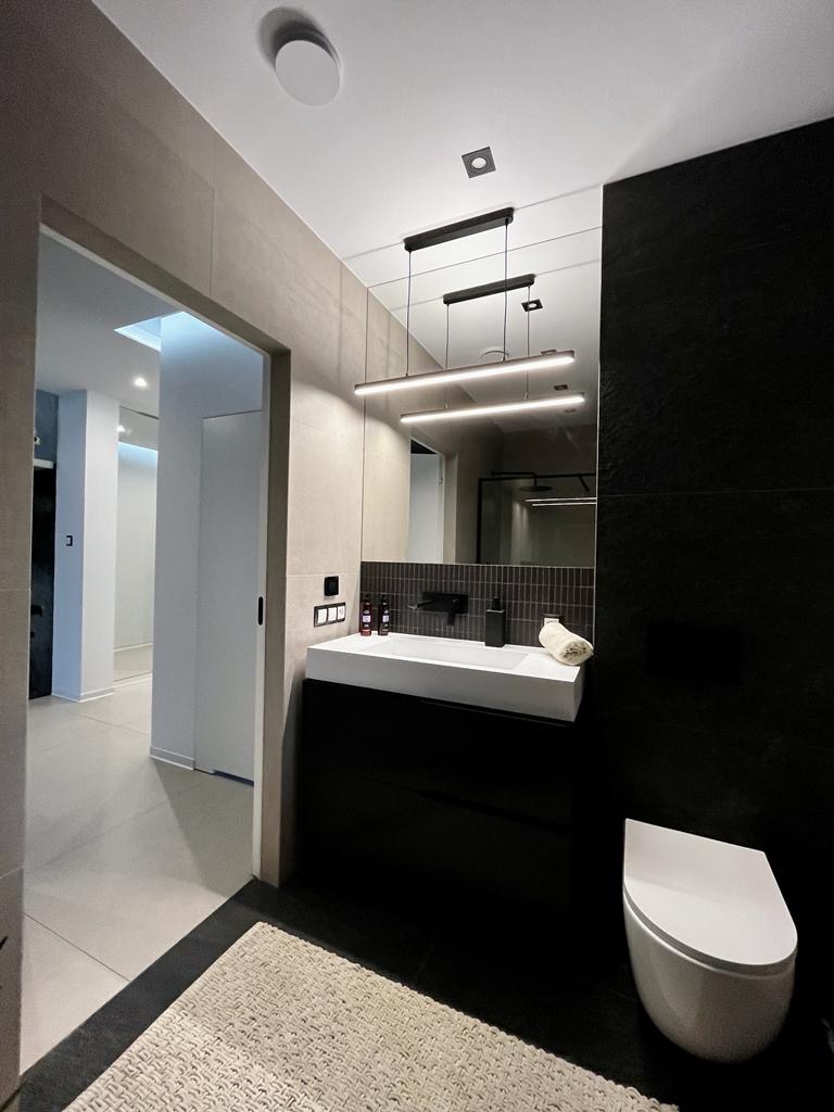 Nowoczesna łazienka z czarną ścianą, podwieszaną umywalką z szafką, wiszącą toaletą i podświetlanym lustrem z trzema lampami