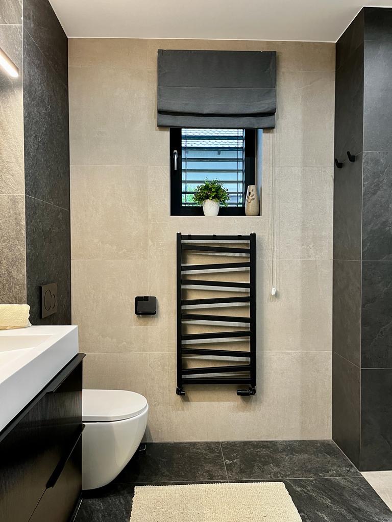 Nowoczesna łazienka z beżowo-szarymi płytkami, czarną armaturą i grzejnikiem drabinkowym na tle okna z roletą rzymską. Na parapecie roślina doniczkowa i ceramiczna figurka kota.