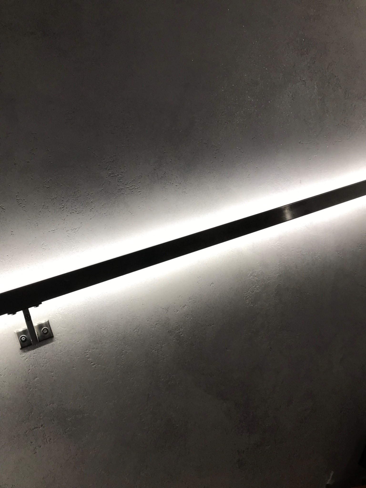Ściana z szarą, chropowatą fakturą i zamontowana poziomo czarna listwa oświetleniowa LED z widocznymi metalowymi mocowaniami.