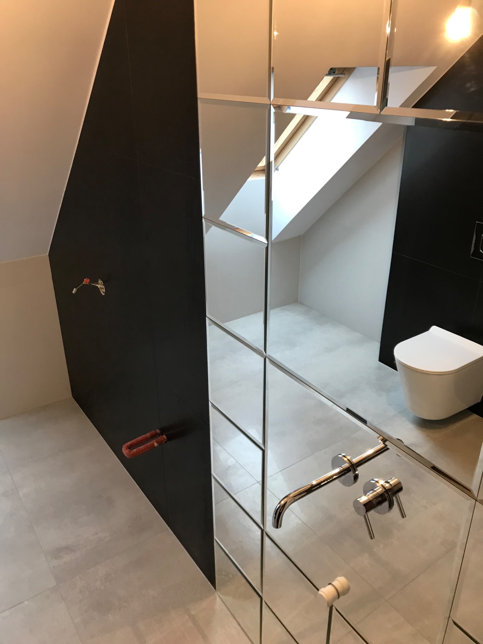 Nowoczesna łazienka na poddaszu z czarno-białymi płytkami, wiszącą toaletą i lustrzaną ścianą z chromowaną armaturą, widok na okno dachowe.