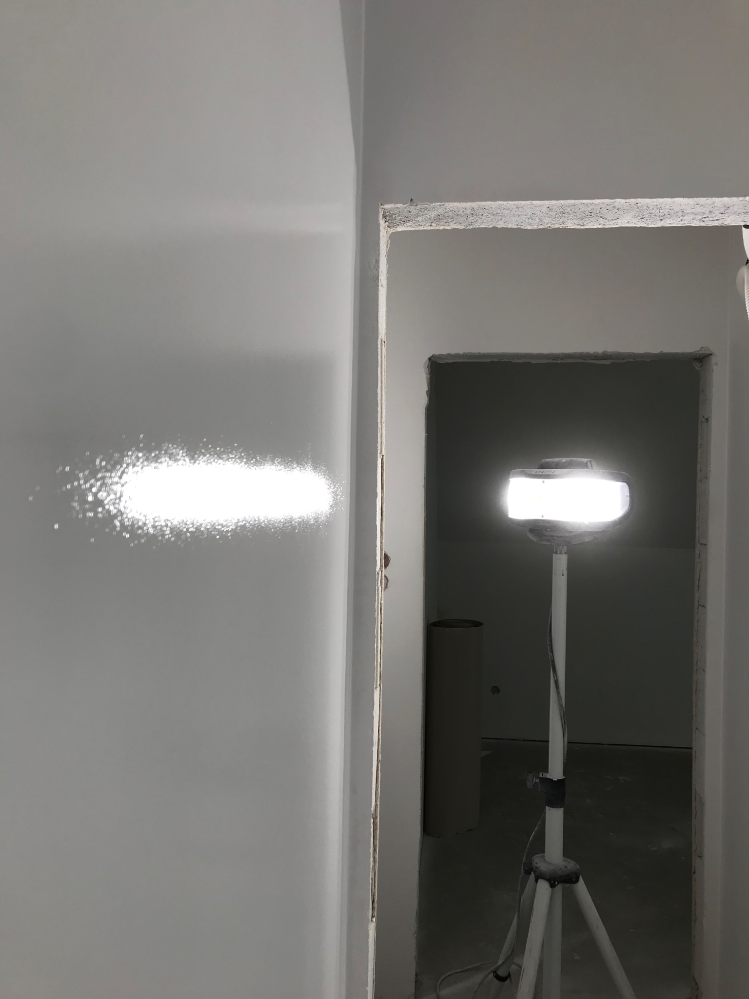 Świeżo pomalowana, gładka ściana oświetlona lampą roboczą na statywie, widoczny niedokończony otwór drzwiowy z surowym wykończeniem.