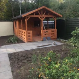 Nowo wybudowana, drewniana altana ogrodowa z ażurowymi ściankami, położona na wyłożonym płytami tarasie, z widocznym ogrodem w trakcie aranżacji.