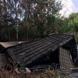 Przeprowadzki Od A do Z - Spalona szopa z czarnymi śladami po pożarze na dachu i ścianach, otoczona zielenią i gruzem.