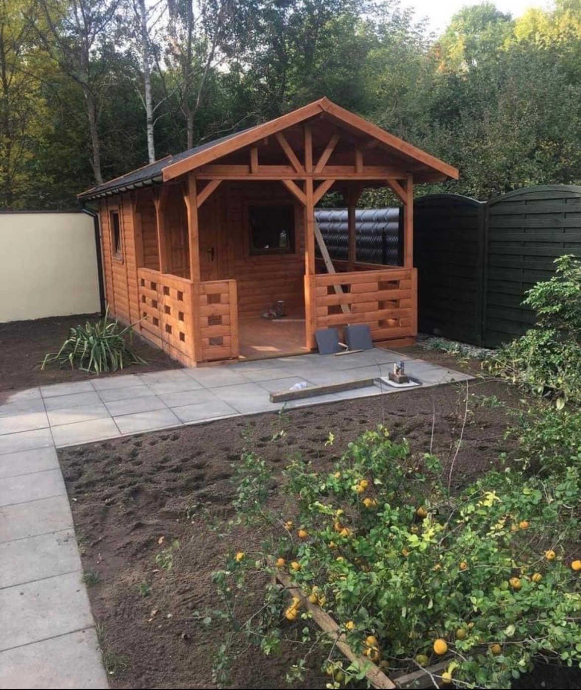 Nowo wybudowana, drewniana altana ogrodowa z ażurowymi ściankami, położona na wyłożonym płytami tarasie, z widocznym ogrodem w trakcie aranżacji.