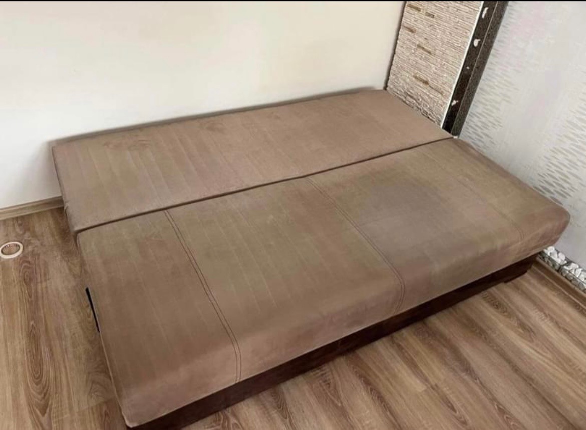 Rozłożona, brązowa kanapa z widocznymi śladami użytkowania na tapicerce, ustawiona na panelach podłogowych w pokoju z jasnymi ścianami.