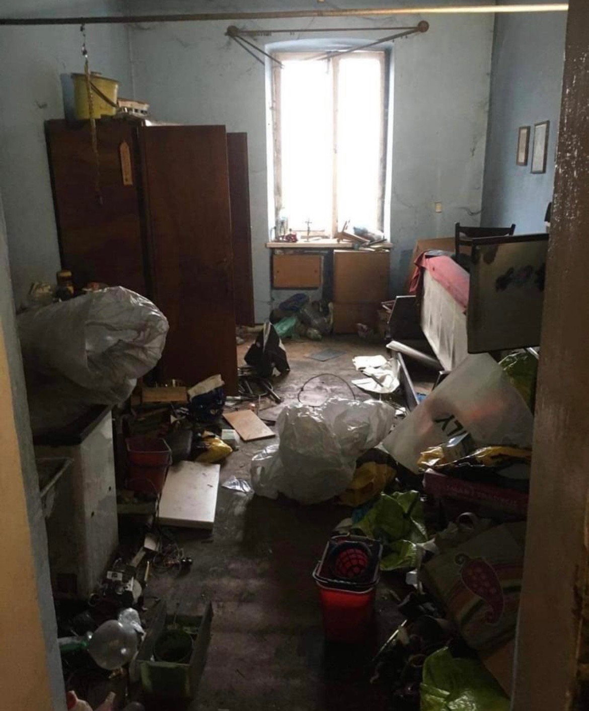 Zrujnowany pokój z zawalonymi śmieciami podłogami, starymi meblami i odpadami w plastikowych workach, widok zza framugi drzwi.