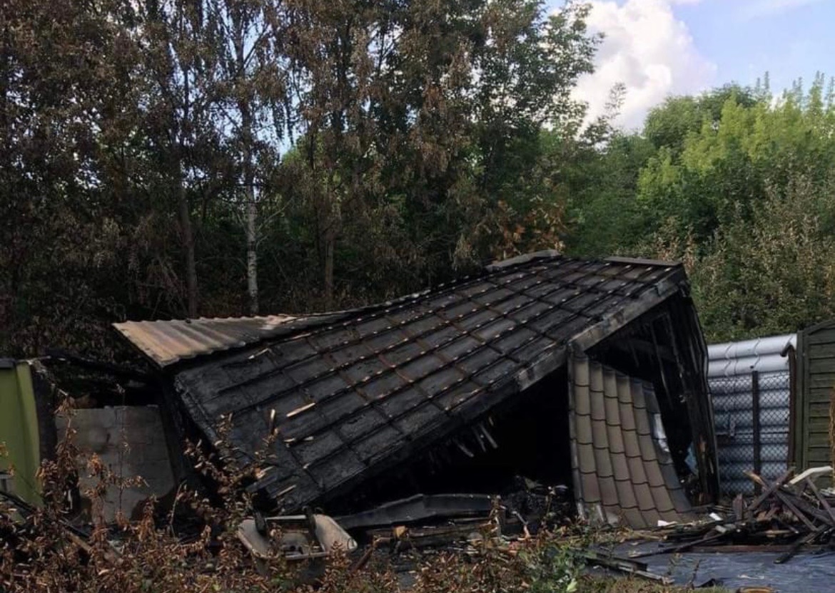 Spalona szopa z czarnymi śladami po pożarze na dachu i ścianach, otoczona zielenią i gruzem.