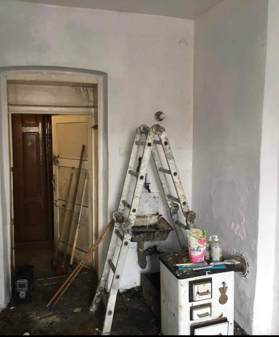 Wnętrze pokoju w trakcie remontu: drabina oparta o ścianę, brudna umywalka, piec kaflowy z płynem do mycia, ślady farby na ścianach i podłodze, otwarte drzwi do kolejnego pomieszczenia.