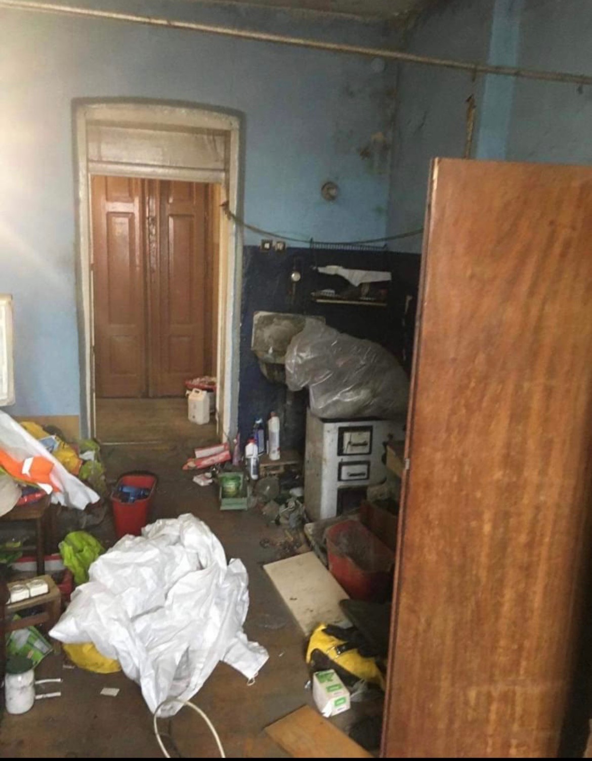 Zaniedbane wnętrze pokoju z widocznymi śladami wilgoci na ścianach, zagracone podłogi i meble, duży bałagan przed drzwiami do kolejnego pomieszczenia.