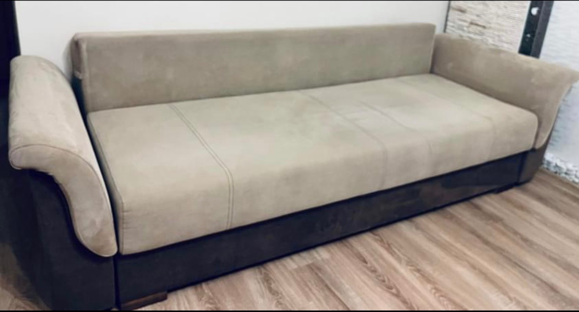 Beżowa sofa z ciemnobrązową podstawą na panelach podłogowych, widok z boku.