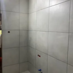 Ściana w łazience wyłożona szarymi, kwadratowymi płytkami, widoczne otwory pod instalacje i wystający zawór.