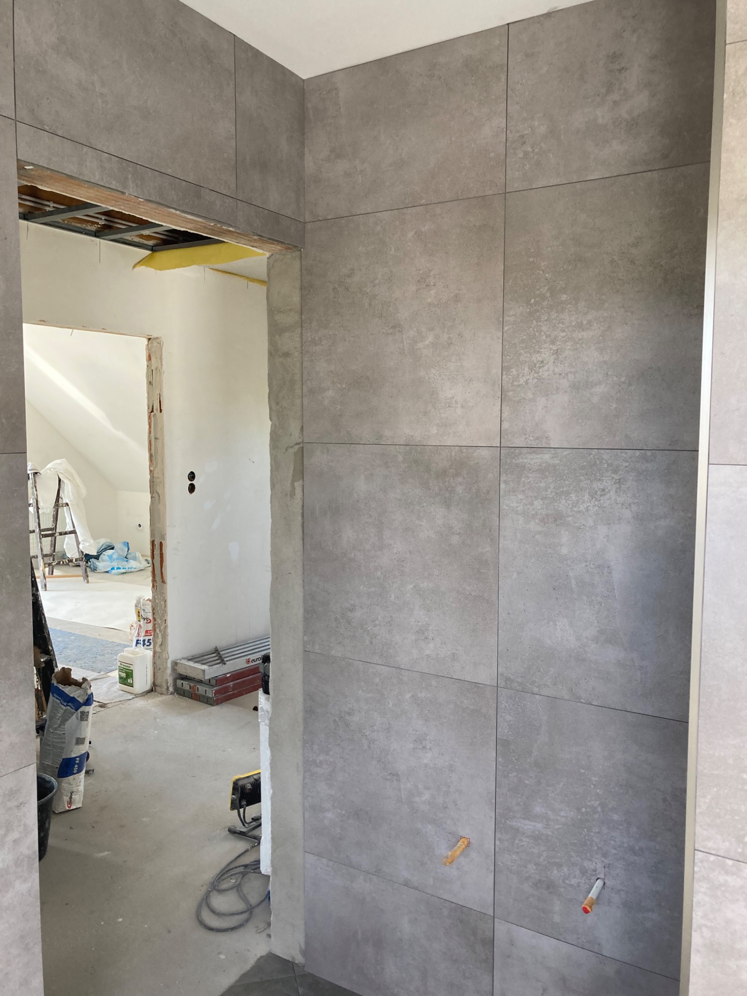 Ściana wyłożona szarymi płytkami w trakcie remontu, widoczne otwory na instalacje i surowe wykończenie futryny drzwiowej prowadzącej do pomieszczenia z drabiną i workami z zaprawą.