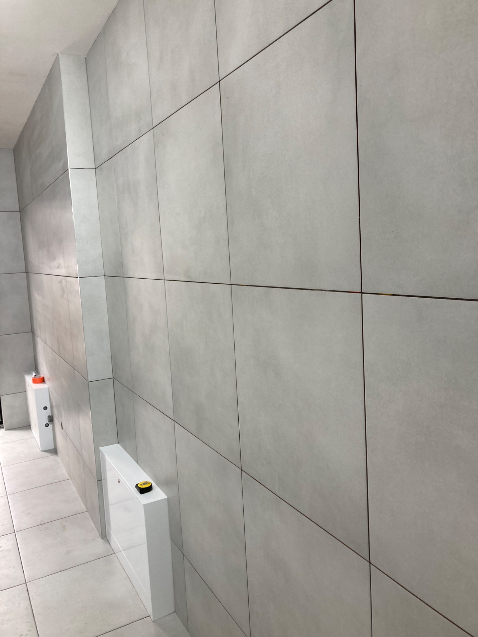 Ściana w łazience wyłożona dużymi, szarymi płytkami ceramicznymi o matowym wykończeniu, widoczne fugi w kolorze ciemnego brązu, biała zabudowa z żółtą poziomicą na górze, ujęcie z perspektywy podłogi.