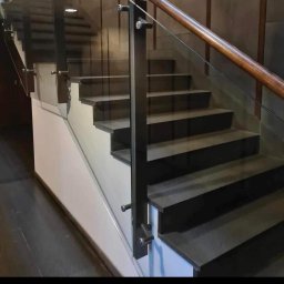 Robik , Sylwia Stępień - Nowoczesne schody z ciemnymi stopniami, szklaną balustradą i drewnianą poręczą, prezentowane w pomieszczeniu z szarymi panelami ściennymi.