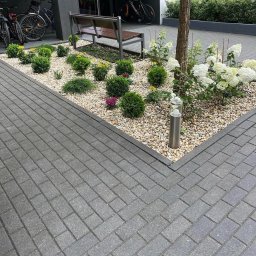 Robik , Sylwia Stępień - Uporządkowany teren przed budynkiem z kostki brukowej, ozdobnym żwirowym klombem z kulistymi krzewami, hortensjami i ławką, rowery zaparkowane w tle.