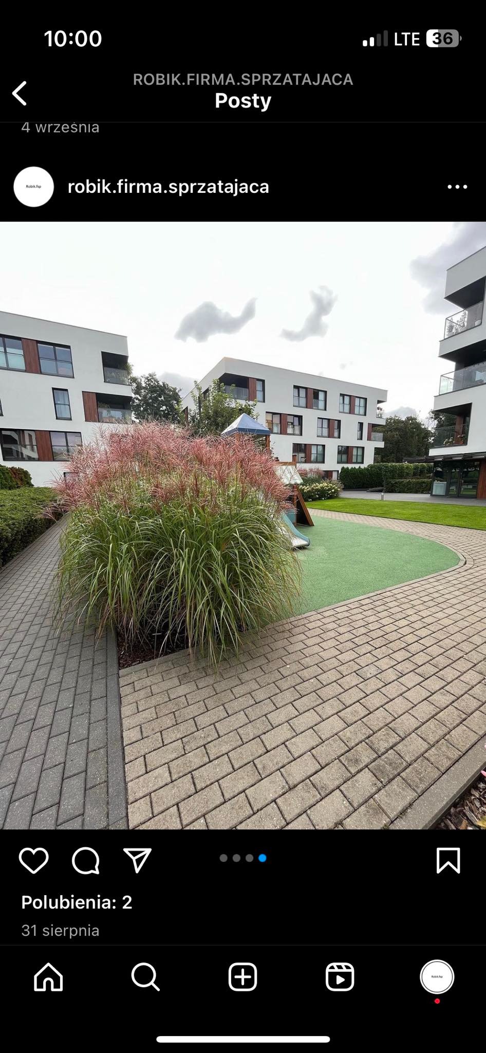 Widok na plac zabaw z zieloną nawierzchnią i niską zjeżdżalnią, otoczony ozdobną trawą i chodnikiem z kostki brukowej, na tle nowoczesnego budynku mieszkalnego.