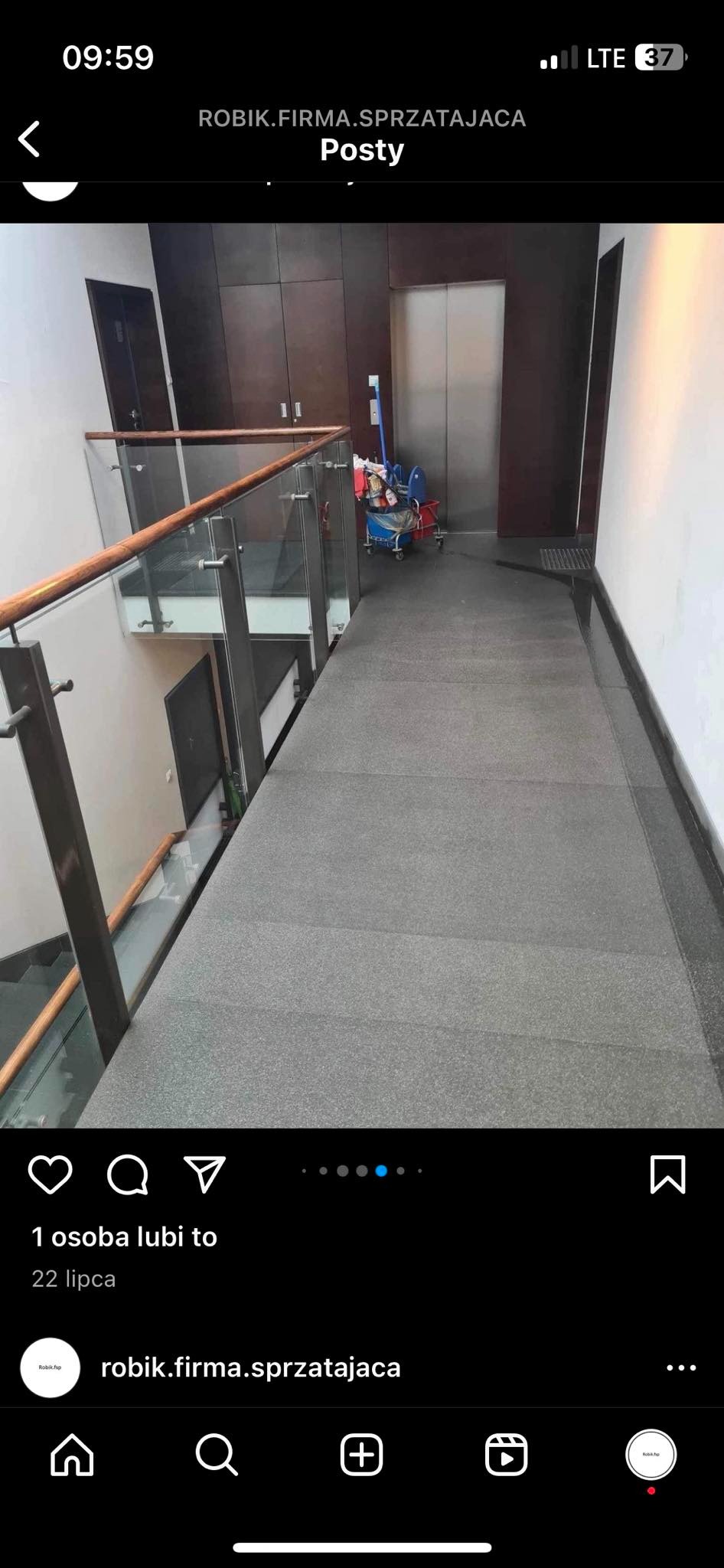 Korytarz w budynku biurowym z szarą wykładziną, szklaną balustradą i wózkiem do sprzątania w tle, stojącym obok windy.