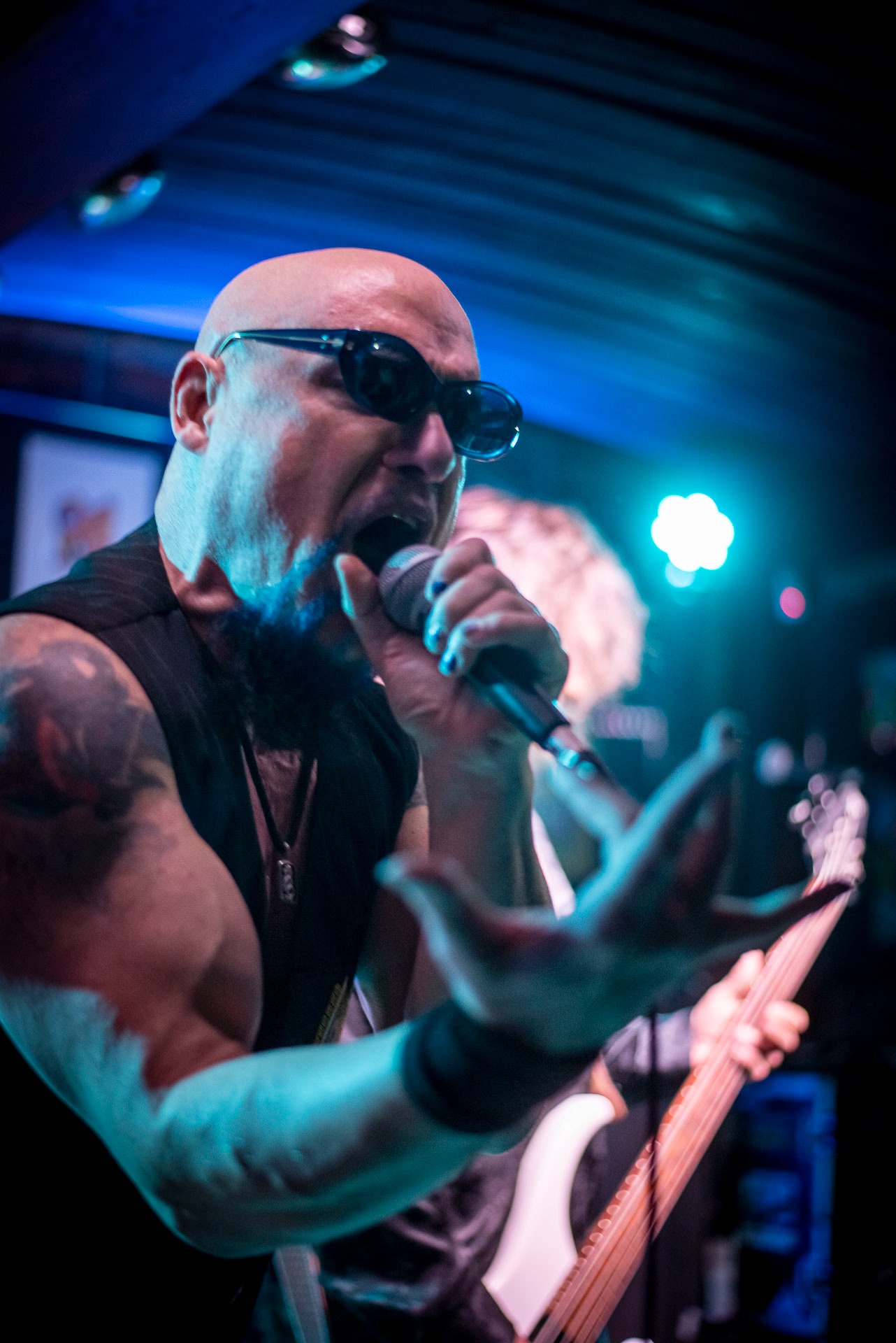 Mężczyzna z ogoloną głową i tatuażem na ramieniu śpiewa do mikrofonu na scenie, w tle widoczny gitarzysta i oświetlenie sceniczne.
