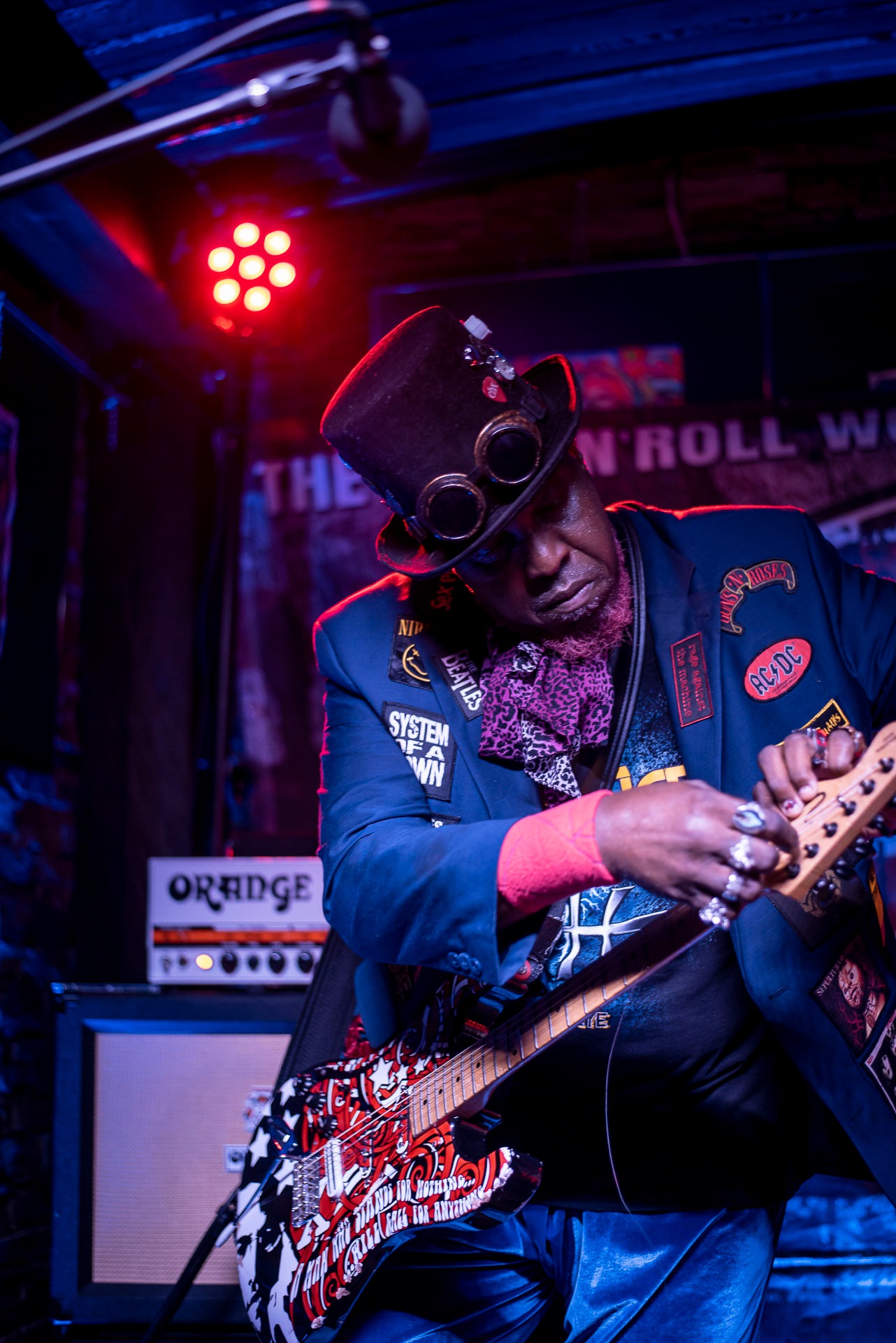 Muzyk w kapeluszu steampunk i niebieskim garniturze z naszywkami gra na elektrycznej gitarze z wzorem w kolorach czerwieni, bieli i czerni na scenie z oświetleniem scenicznym i wzmacniaczem Orange...