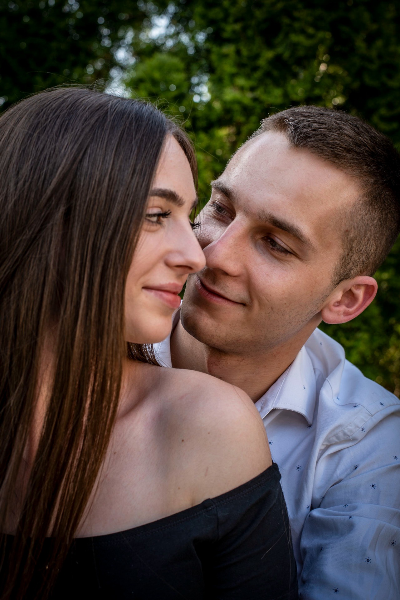 Młoda para w bliskim ujęciu, ona w czarnej sukience z odkrytymi ramionami, on w białej koszuli, oboje patrzą na siebie z uśmiechem, tło z zielenią.