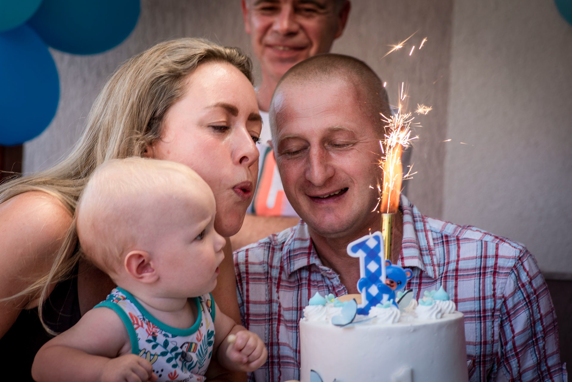 Rodzina świętuje pierwsze urodziny dziecka: mama zdmuchuje świeczkę, tata uśmiecha się z zamkniętymi oczami, a dziecko patrzy z zaciekawieniem na tort z cyfrą jeden i płonącą fontannę.
