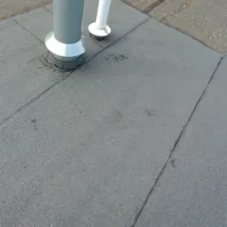 Szary dach pokryty papą z widocznymi kominami wentylacyjnymi, zgrzewami papy i butelką wody w tle.