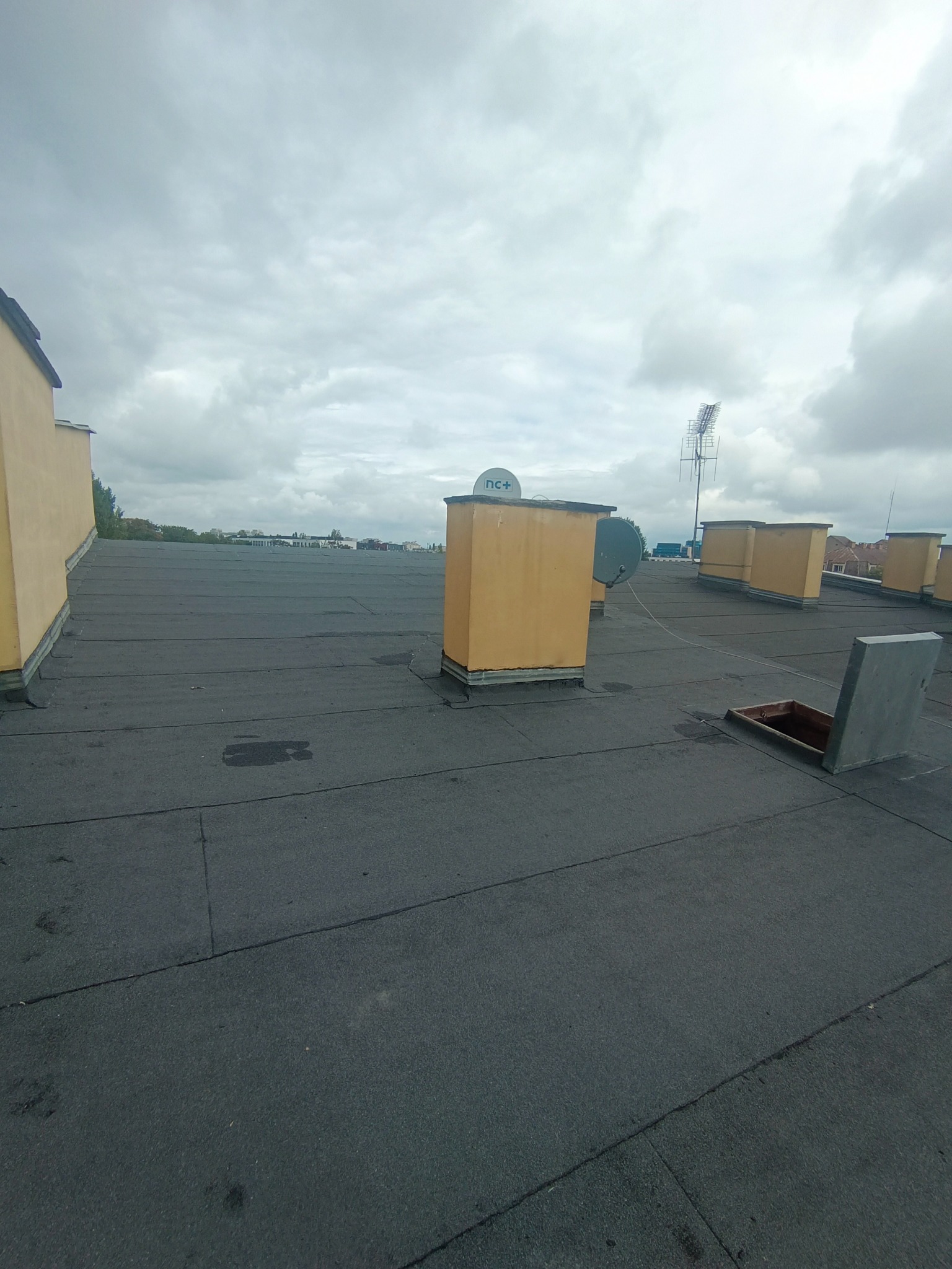 Czarny, płaski dach pokryty papą z kominami wentylacyjnymi, anteną satelitarną i otwartym włazem na tle pochmurnego nieba.