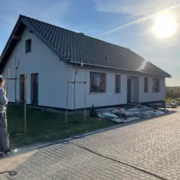 Świeżo otynkowany dom jednorodzinny z ciemną dachówką, okna zabezpieczone folią, przygotowany do dalszych prac wykończeniowych, widoczne materiały budowlane na palecie obok budynku.