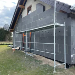 Dom w trakcie izolacji termicznej ścian zewnętrznych, widoczne rusztowanie i czarne płyty styropianowe z szarymi kołkami mocującymi, częściowo otynkowana ściana szczytowa