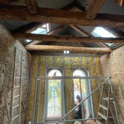 La-Bud usługi budowlane - Wnętrze poddasza z odsłoniętą cegłą i drewnianymi belkami stropowymi, ocieplone wełną mineralną w metalowym stelażu, widoczne okna dachowe i drabiny.
