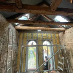 Wnętrze poddasza z odsłoniętą cegłą i drewnianymi belkami stropowymi, ocieplone wełną mineralną w metalowym stelażu, widoczne okna dachowe i drabiny.