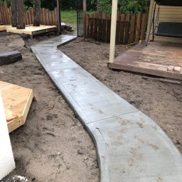 La-Bud usługi budowlane - Świeżo wylana, betonowa ścieżka wijąca się przez ogród, prowadząca od drewnianego tarasu z huśtawką do metalowej bramki z drewnianym ogrodzeniem w tle. Wokół ścieżki widoczna świeżo rozkopana ziemia.