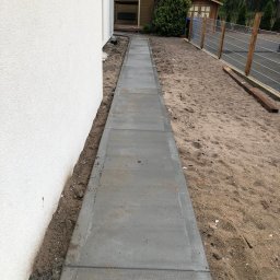 La-Bud usługi budowlane - Świeżo ułożony, betonowy chodnik z szarych płyt prowadzący wzdłuż białej ściany budynku, z widocznym piaskiem i ziemią po bokach oraz fragmentem ogrodu z drzewami w tle.