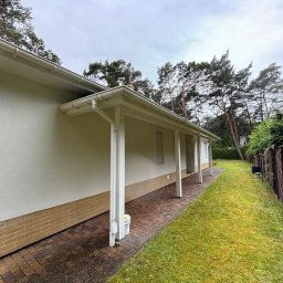 La-Bud usługi budowlane - Jasny dom jednorodzinny z białymi kolumnami werandy i elewacją w odcieniach beżu, widok z boku na tle zielonego trawnika i drzew